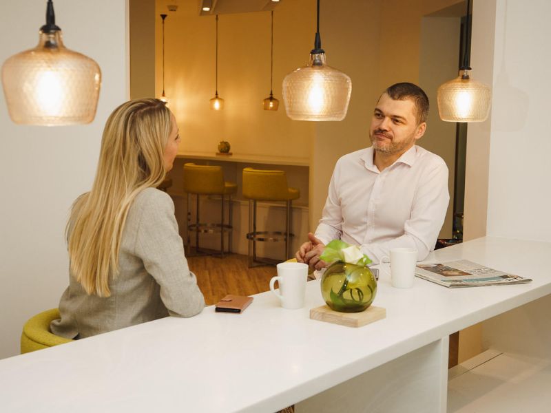 A female and male sitting at a bench style worktop, talking beneath ambient lighting.