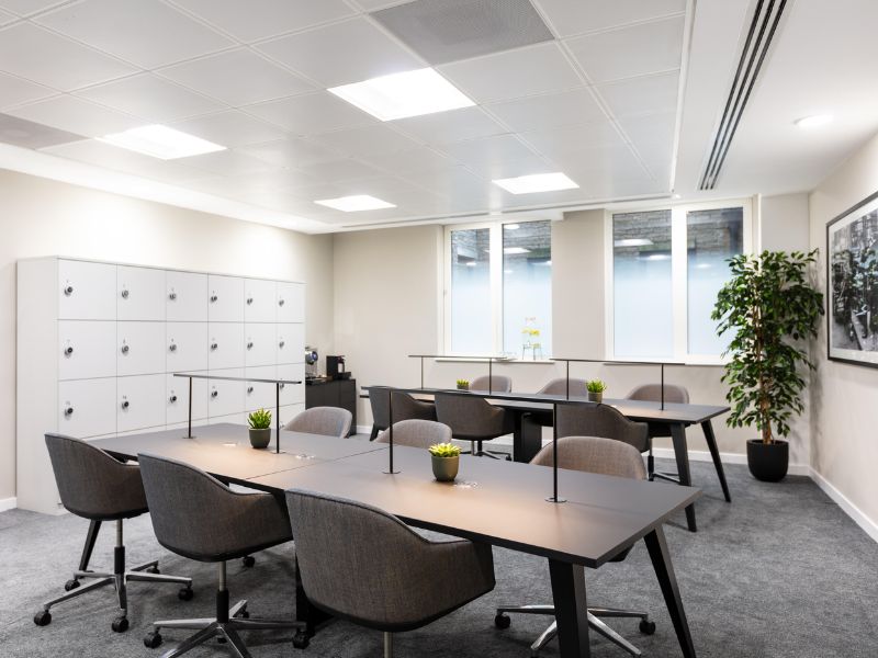 A coworking space with two tables, six chair on each with lockers in the background.